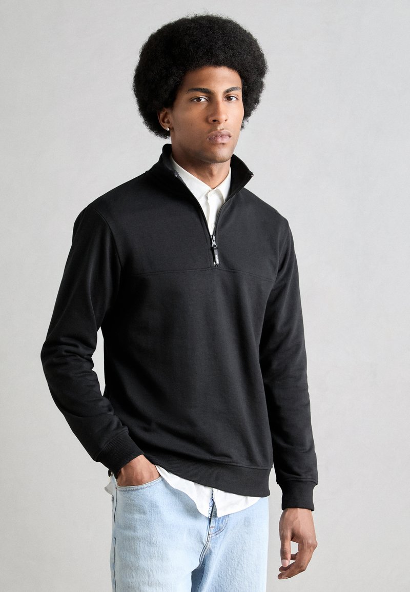 Black pullover sweatshirt with a quarter-zip collar, ribbed cuffs, and a smooth texture, worn over a white collared shirt.
