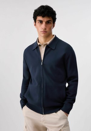 Joven con chaqueta azul marino con cremallera sobre una camisa polo beige y pantalones cargo beige, de pie con las manos en los bolsillos.