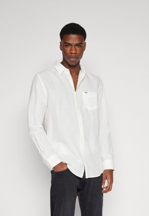 Camicia di lino bianca con colletto a punta e chiusura con bottoni, dotata di una singola tasca sul petto e branding discreto. Indossata con pantaloni scuri.