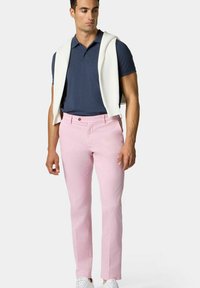 Chinos rosa chiaro in cotone, con vestibilità slim, chiusura a bottone in vita e tasche laterali. Abbinati a un polo blu navy e a un maglione bianco drappeggiato sulle spalle.