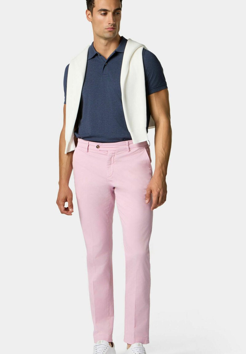 Chinos rosa chiaro in cotone, con vestibilità slim, chiusura a bottone in vita e tasche laterali. Abbinati a un polo blu navy e a un maglione bianco drappeggiato sulle spalle.