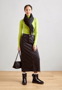 Grön långärmad polotröja, svarts denim maxikjol, svart fransad scarf, svarta ankelskor och en liten svart handväska. Släta texturer, avslappnad stil.