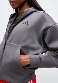 Άτομο που φοράει γκρι hoodie με φερμουάρ και μπροστινή τσέπη με λογότυπο, χέρι μέσα στην τσέπη, σε συνδυασμό με παντελόνια με κόκκινες και λευκές ρίγες.