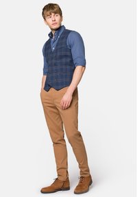 Granatowa marynarka w kratę założona na jasnoniebieską koszulę, połączona z jasnobrązowymi, dopasowanymi spodniami i brązowymi zamszowymi butami. Warto zwrócić uwagę na charakterystyczne guziki na kamizelce.