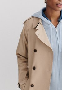 Beige Trenchcoat mit großen braunen Knöpfen, gerippten Manschetten und einem breiten Kragen, über einem hellblauen Hoodie mit Reißverschluss und Kordeln.