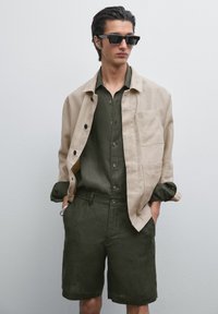 Beige linen jacket with black buttons, green linen shirt, and matching shorts. Model wearing black sunglasses, arms relaxed at sides.