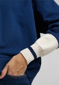Sudadera azul marino con puños de crema acanalados que presentan dos rayas en azul marino. Textura de tela suave, corte relajado y detalle de bolsillo lateral visible.