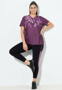Paarse actieve t-shirt met bloemenontwerp, gecombineerd met zwarte leggings met een roze accentstreep. Zwarte sportieve schoenen maken de outfit compleet.