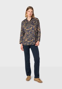 Flowy, patterned blouse in navy and gold, featuring a collar and long sleeves, paired with dark trousers and light brown shoes.