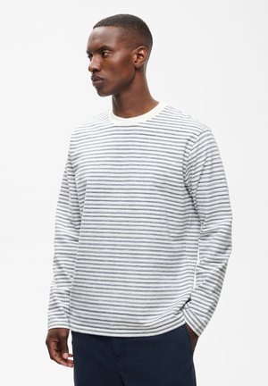 Mann trägt ein weißes und graues gestreiftes Langarmshirt und dunkle Hose, blickt nach links mit einer Hand in der Tasche.