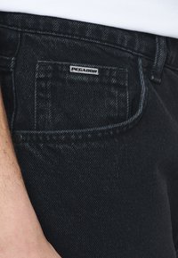 Vaqueros de mezclilla negros con un bolsillo trasero visible. Presenta una etiqueta de la marca "PEGADOR" cosida en el bolsillo. Textura lisa con un ligero brillo.