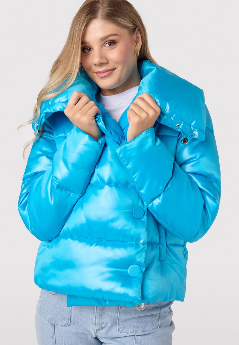 Helderblauwe pufferjack met een grote kraag, glanzende afwerking en oversized knopen. Het model houdt de jas gesloten.