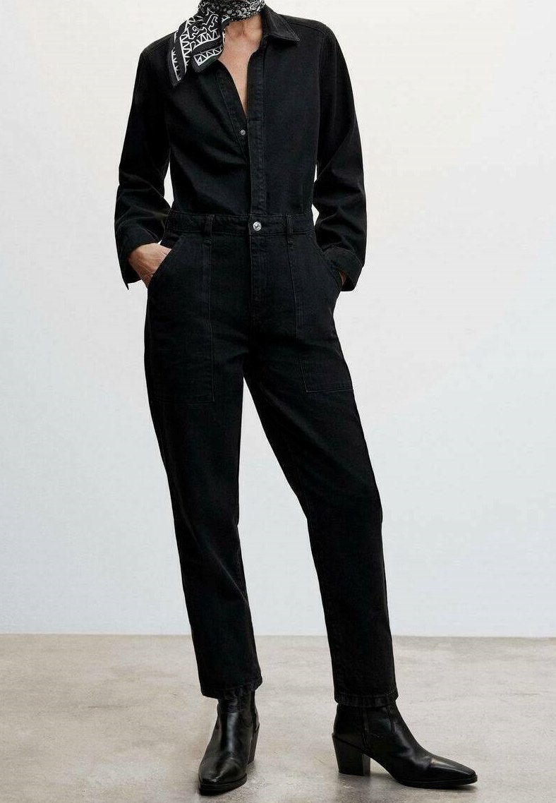 Personne portant une combinaison noire à manches longues avec des poches, des bottines noires à talons et un foulard noir et blanc à motifs autour du cou.