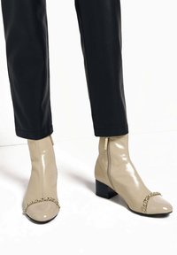 Béžové lakované polokozačky s řetízkovým detailem na špičce, se silnými podpatky a bočním zipem, v kombinaci s černými kalhotami.