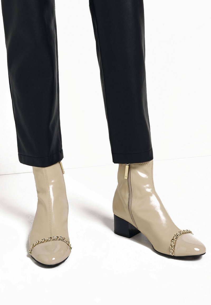 Béžové lakované polokozačky s řetízkovým detailem na špičce, se silnými podpatky a bočním zipem, v kombinaci s černými kalhotami.