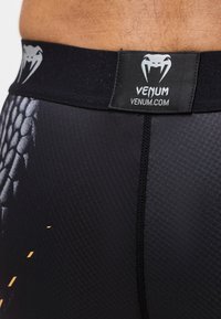 Κοντινό πλάνο στη ζώνη μαύρου αθλητικού ρούχου Venum με λευκό λογότυπο φίδι και σχέδιο φολίδων φιδιού στο ύφασμα κοντά στη μέση.