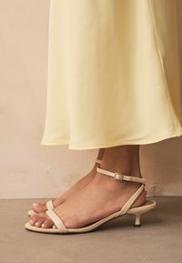 Sandales beiges à brides avec une finition texturée, fines sangles à la cheville, et un petit talon kitten, portées avec une robe jaune fluide.