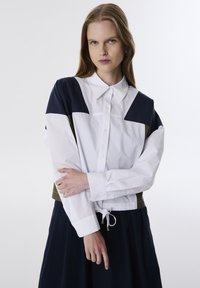 Chemise à col blanc avec des accents bleu marine et olive, dotée d'une taille à cordon et de manches longues. Tissu lisse, design structuré.