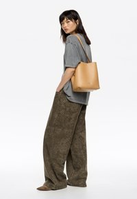 Borsa a secchiello in cuoio marrone chiaro con finitura liscia e rivetti metallici, indossata sulla spalla, abbinata a pantaloni larghi verdi oliva con motivo e una maglietta grigia.