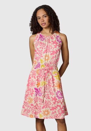 Kvinne med krøllete hår iført en ermeløs rosa kjole med oransje, lilla og gule blomster mønstre som står mot en ensfarget bakgrunn.