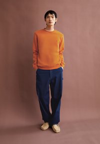 Maglione girocollo arancione, pantaloni a gamba larga blu navy, scarpe slip-on beige. Design minimalista, texture liscia, logo ricamato sul petto.
