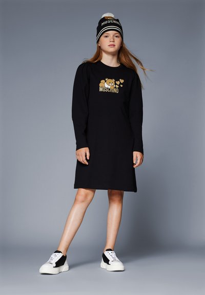 Abito lungo nero a maniche lunghe con grafica di un orso dorato e testo. Abbinato a sneakers bianche e nere. Modella con un berretto bianconero.