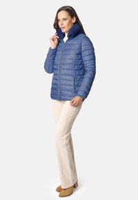 Chaqueta de plumas azul con cremallera frontal y cuello de tejido azul marino. Presenta costuras horizontales y bolsillos laterales. Combinada con pantalones beige y botas marrón claro.