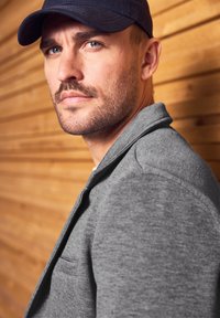 Harmaa fleece-takki, jossa on kaulus ja sivutasku. Henkilöllä on päässään tummansininen baseball-lippalakki puutaustalla.