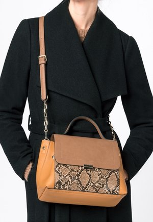 Frau mit schwarzem Mantel, die eine Tasche aus braunem Leder mit Schlangenprint und einem oberen Griff sowie einem Schultergurt trägt.