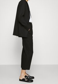 Zwarte op maat gemaakte colbert, strakke zwarte broek en zwarte leren muiltjes met zilveren hardware. Minimalistisch ontwerp met een strakke silhouet.