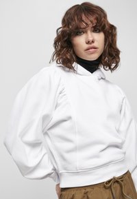 Felpa bianca con maniche a sbuffo e orlo a costine; presenta un grande cappuccio e cuciture strutturate. Abbinata a pantaloni beige con coulisse.