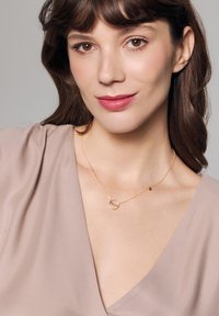 Złoty naszyjnik z okrągłym wisiorkiem i małym zawieszką, noszony na jasnobeżowej bluzce z dekoltem w kształcie litery V. Gładka tekstura i delikatny design.