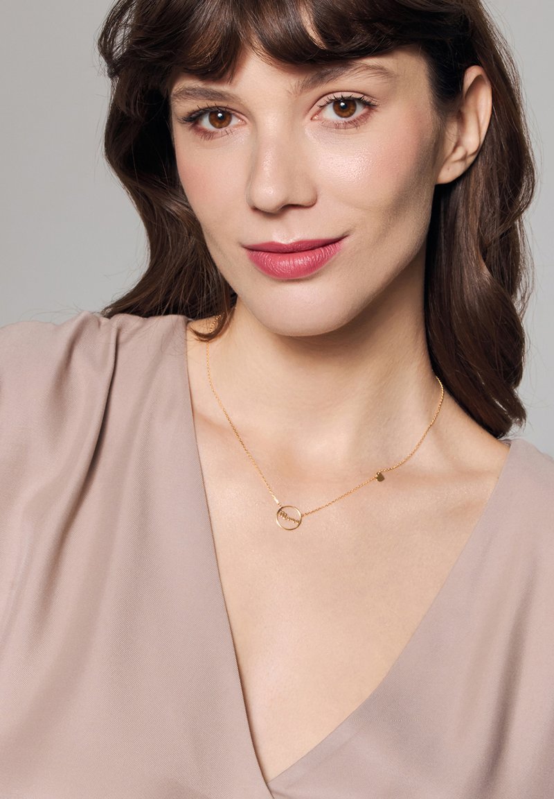 Złoty naszyjnik z okrągłym wisiorkiem i małym zawieszką, noszony na jasnobeżowej bluzce z dekoltem w kształcie litery V. Gładka tekstura i delikatny design.