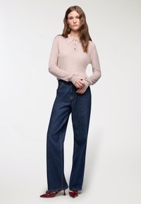 Kobieta stoi w jasnoróżowej bluzce z długim rękawem, ciemnoniebieskich szerokich dżinsach i czerwonych szpilkach na białym tle.