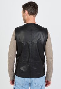 Schwarze Lederweste mit glatter Textur, ohne Nähte am Rücken, getragen über einem hellgrauen Langarmshirt und blauen Jeans.