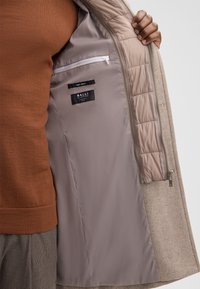 Persoon die een beige wollen jas openhoudt, waarbij de lichtgrijze binnenvoering met een ritszak en labels zichtbaar is.