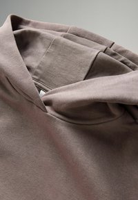 Sudadera marrón con un tejido suave y texturizado. Presenta una gran capucha, costuras cosidas y una pequeña etiqueta blanca en el interior. Diseño sencillo y funcional.