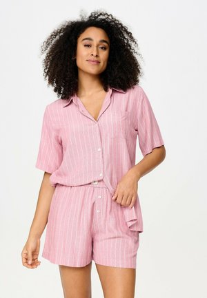 Femme aux cheveux bouclés portant une chemise rose à manches courtes boutonnée et un short assorti avec des rayures verticales blanches sur un fond uni.