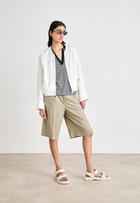 White bomber jacket, striped black and white top, beige shorts, and light-colored sandals. Accessories include gray sunglasses. Smooth textures.