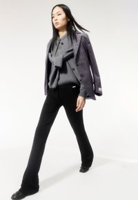 Grauer Pullover mit Schleifendetail, lila Cordblazer, schwarze Hose und schwarze Schuhe. Glatte Texturen und maßgeschneiderte Passformen werden hervorgehoben.