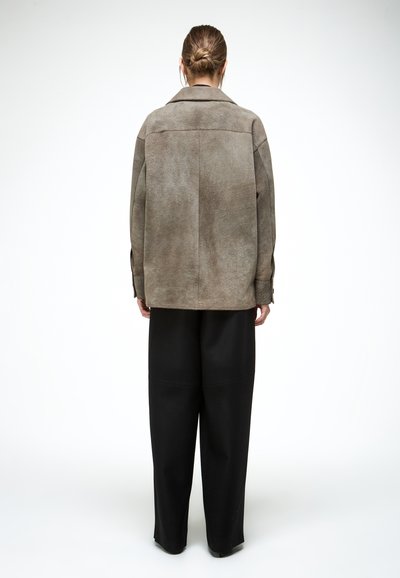 Osoba s vlasmi zviazanými vzadu, ktorá má na sebe voľnú taupe semišovú bundu a široké čierne nohavice, stojaca chrbtom k divákovi na bielom pozadí.