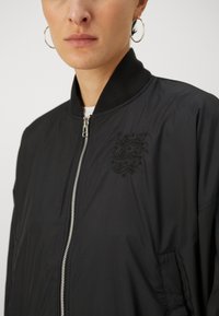 Black bomber jacket made of smooth fabric, featuring a zip front, ribbed collar, and an embroidered design on the left chest.