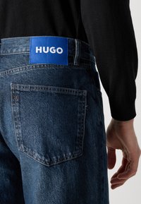 Tumedat siniseid teksaseid, millel on nähtav tagatasku ja sinine nahkpatch, millel on kirjas "HUGO", vöökoha juures. Sile tekstuur.