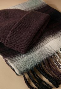 Dark purple knitted beanie atop a plaid scarf in shades of gray and purple, featuring fringed edges and a soft texture.