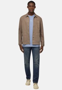Giacca di tela marrone con chiusura a bottoni e due tasche, indossata sopra una maglietta azzurra chiara, abbinata a jeans blu scuro e scarpe beige.