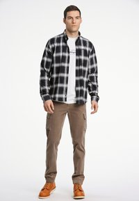 Camicia flanella a quadri in bianco e nero sopra una maglietta bianca, abbinata a pantaloni cargo khaki e stivali arancioni. L'outfit presenta un design con bottoni.