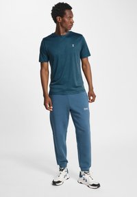 T-shirt bleu marine à manches courtes avec un logo blanc simple, associé à un pantalon de jogging assorti avec poches latérales et poignets élastiques. Chaussures de sport blanches.