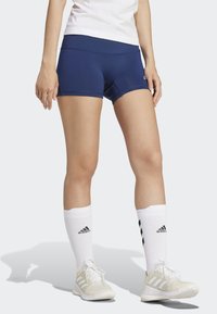 Shorts sportivi blu navy con un design aderente, abbinati a calze bianche al ginocchio con accenti neri del logo e sneakers di colore chiaro.