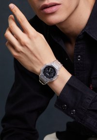 Montre transparente avec un cadran noir et des accents argentés, présentant un affichage numérique et un bracelet transparent ajustable.