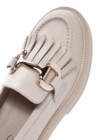 Loafer en cuir beige clair avec une semelle plate, présente des détails en frange et un accent en rose doré sur la sangle supérieure.